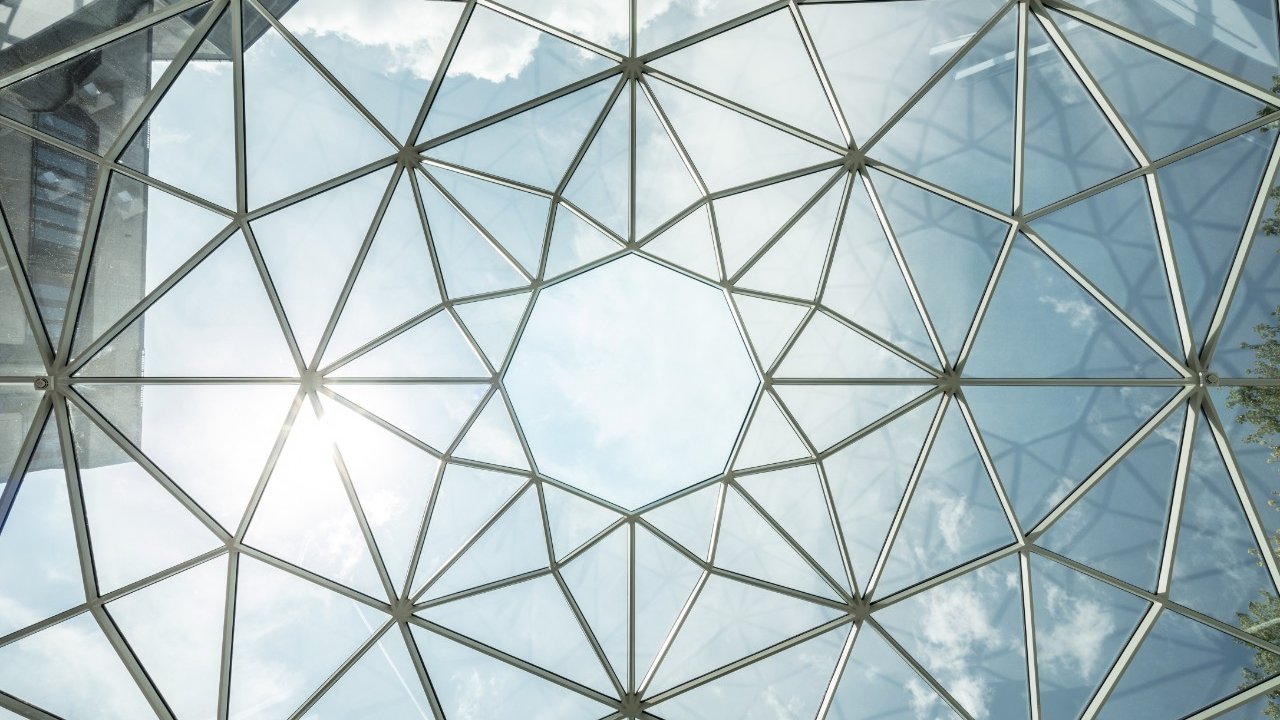 A geometric glass ceiling with triangular patterns, reflecting sunlight and clouds.