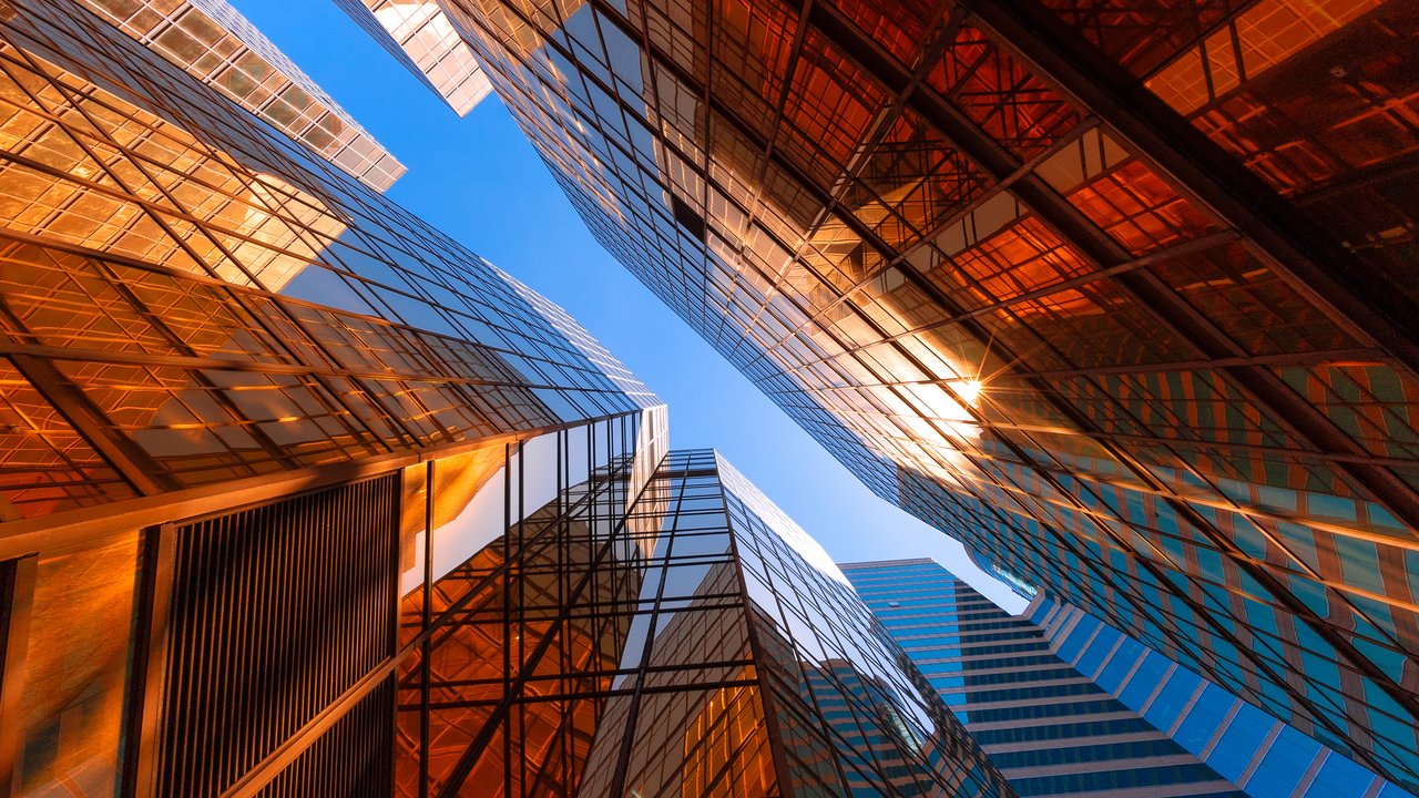 View looking up at tall glass buildings reflecting sunlight against a clear blue sky.