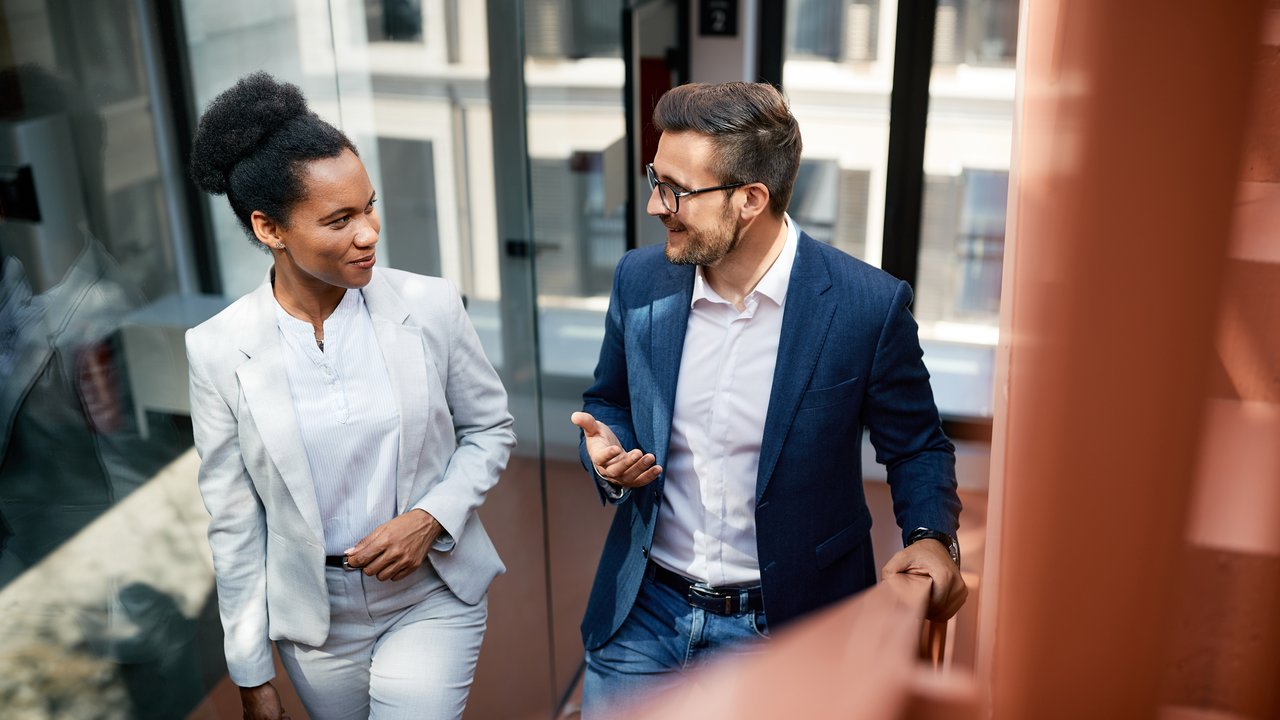 High angle view of a male and a female colleague are talking and smiling while walking up the stairs in a modern office building.