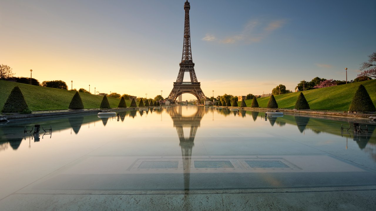 The Eiffel Tower stands tall over a reflective pool, surrounded by manicured lawns and trees at sunrise.
