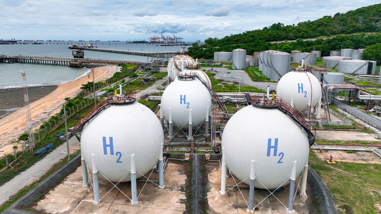 Slightly elevated view of a hydrogen storage facility with industrial H2 gas tanks, wooded hills and sea with a port in the far back.