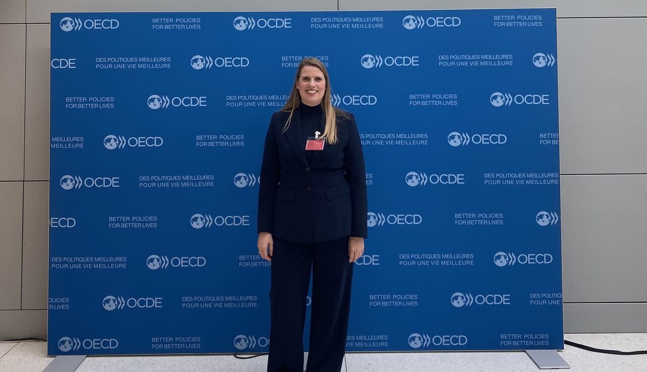 A person in formal attire stands in front of an OECD-branded backdrop with text in English and French.