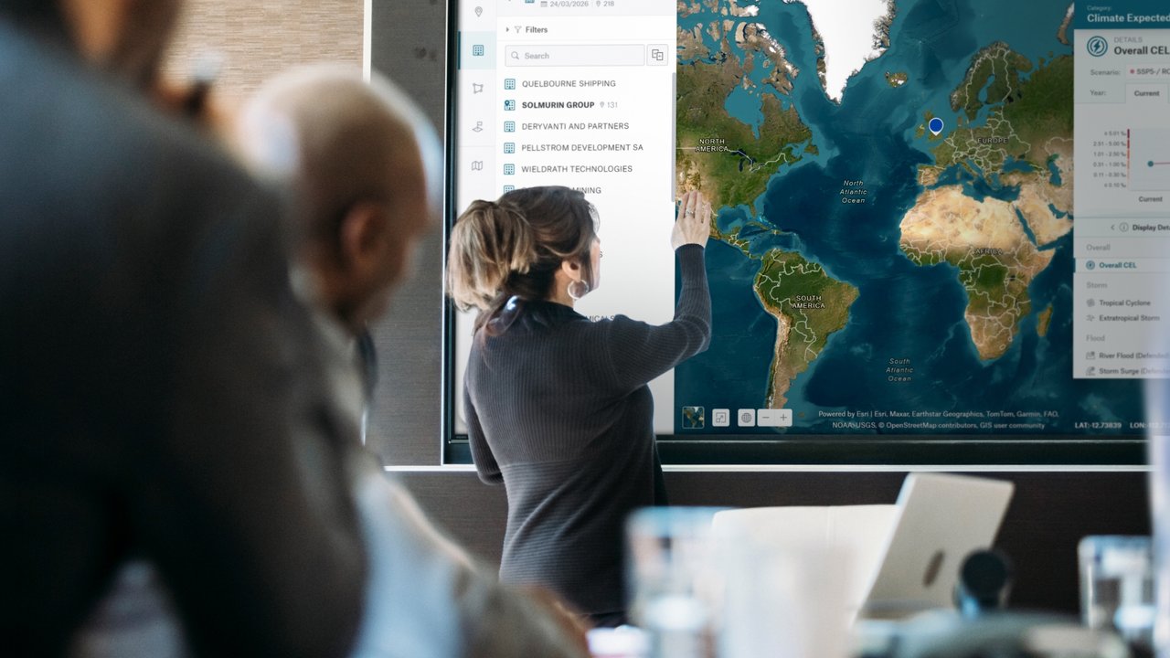 A woman points at a world map displayed on a screen in a meeting room, with blurred figures in the foreground.