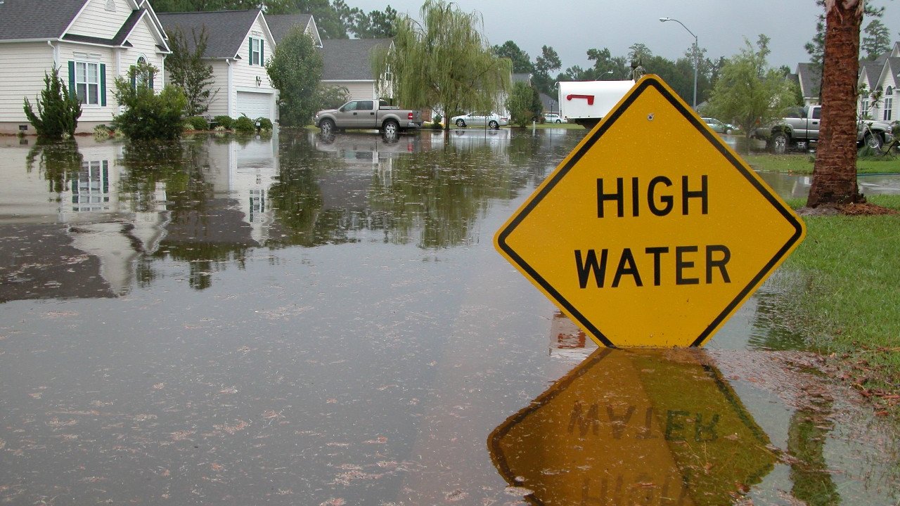 A flooded street with houses in the background and a "HIGH WATER" warning sign in the foreground.
