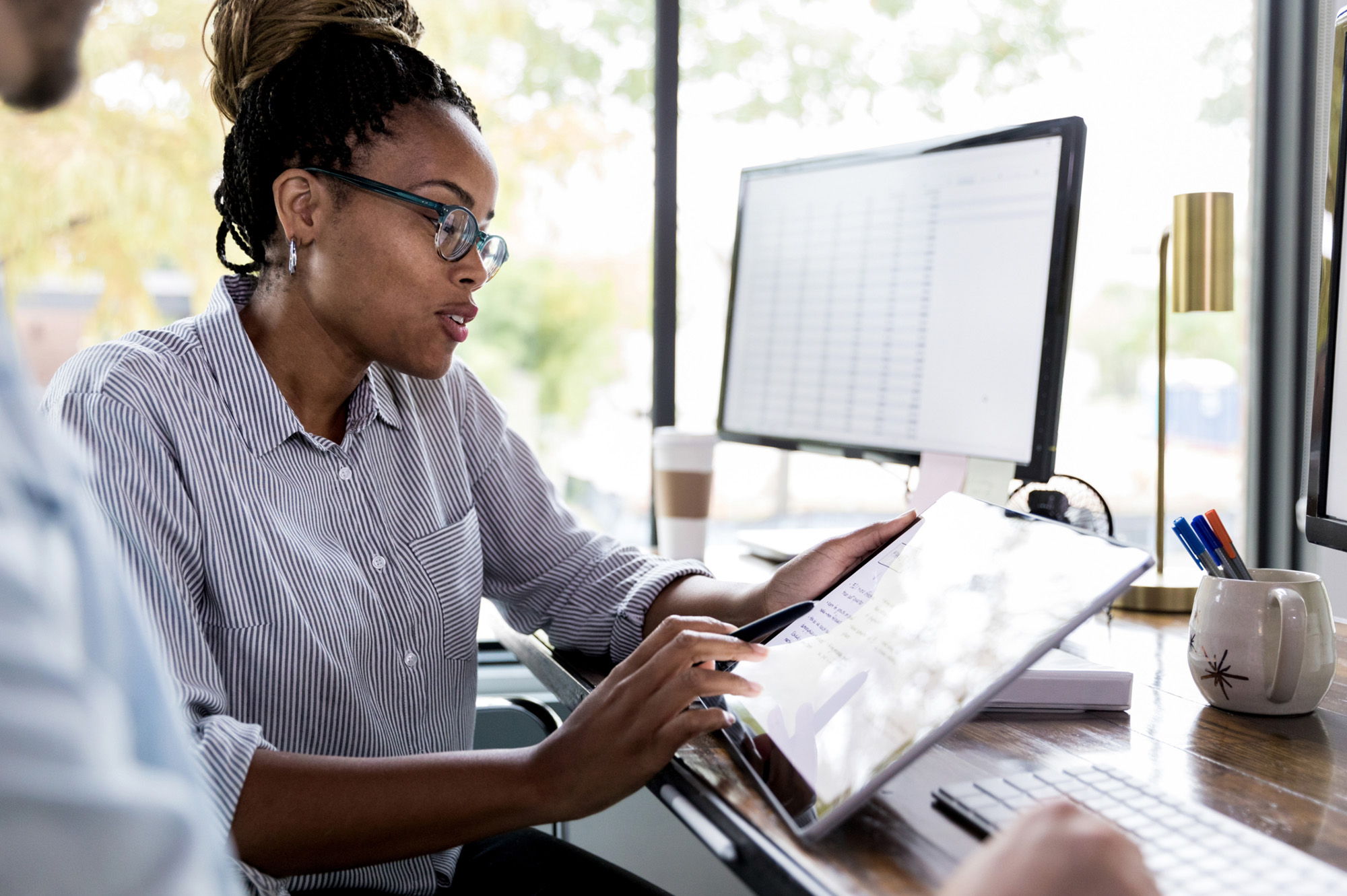 Side view of a woman sitting at an office desk with a co-worker, showing something on a  digital tablet.